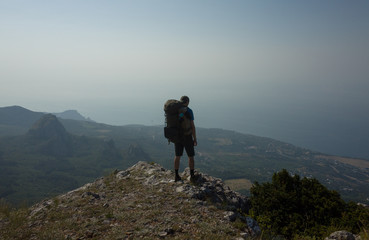 A young tourist with a backpack stands at the edge of the cliff and looks at the mountain landscape and the sea.