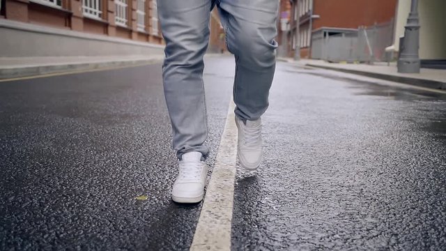A young man with dark hair and a beard, dressed in white sneakers, walks on city road at the dividing white line. The camera moves upwards.