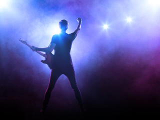 Fototapeta na wymiar Silhouette of guitar player on stage on blue background with smoke and spotlights 