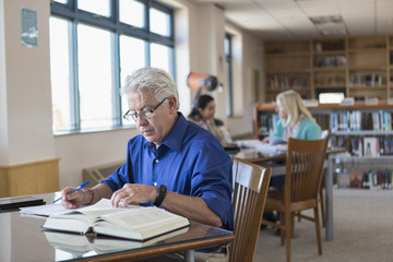 Older man reading book in library and writing notes