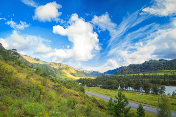 Landscape of the Altai mountains on a summer day. An automobile road that goes to the distance into the mountains between the green coniferous forest, the blue sky with clouds in the mountains