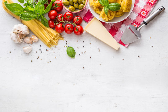 Italian food cuisine and ingredients on white concrete table. Spaghetti Tagliatelle olives olive oil tomatoes parmesan cheese garlic pepper and basil leaves and checkered tablecloth. 