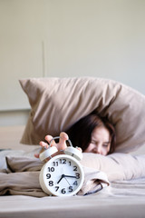 Young beautiful women wake up hurry and extending hand to turn off clock that alarm on morning from under pillow on her bed , focus on clock, soft focus background