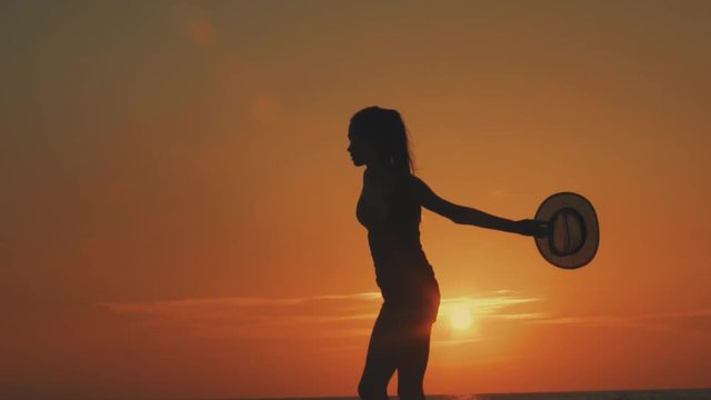 The girl is whirling counterclockwise at sunset or at dawn in Cyprus near the sea, in the distance one sees the horizon and the orange sun, she is holding a straw hat in her hand, slow motion video
