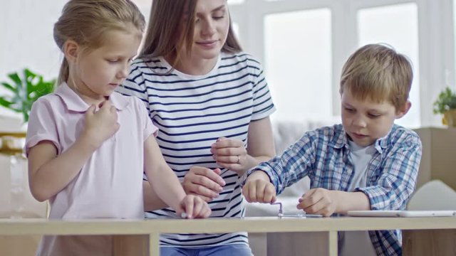 Handheld shot with slow motion of young woman helping little boy with blond hair screwing bolts into furniture while little girl watching and learning