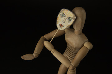 Wooden doll on hinges holds a mask in hands and covers her face on a black background