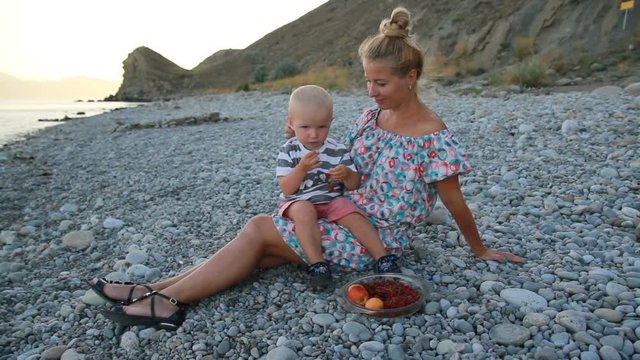 Evening, sea beach. The son sits on his mother's lap and eats fruit and berries