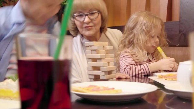 Little boy laughing and discussing jenga tower with grandmother while his cute sister drawing with pencil on table in the cafe