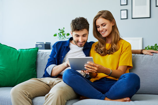 Front View Of A Cute Young Couple Sitting Together On The Couch And Looking At A Tablet