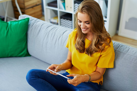 Cheerful Gorgeous Young Lady Sitting On The Sofa At Home And Smiling While Holding Tablet