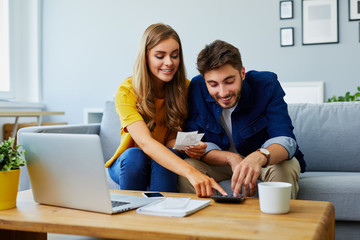 Happy young couple paying bills together and managing budget, sitting on the sofa and using...