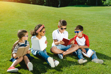 Adorable multiethnic kids in sunglasses reading books on green meadow in park