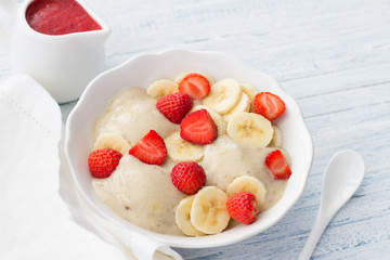 Vegan banana ice cream with strawberry sauce and fresh strawberries in a white bowl on a light blue wooden background