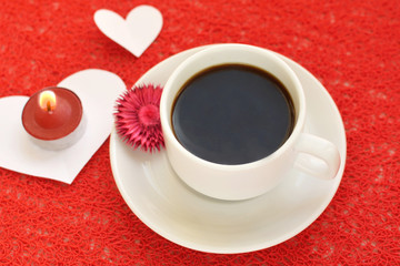 Mug of hot black coffee in a white porcelain mug, card in the shape of heart and a candle on a red lace background