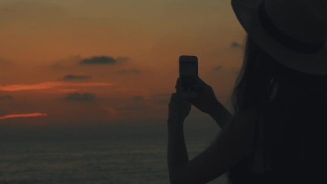 Sexy young woman traveler with a backpack and straw hat on her head takes pictures on mobilephone of sunset and dawn on the sea in Cyprus, orange sun and sky, Silhouette of a girl, close up, 100