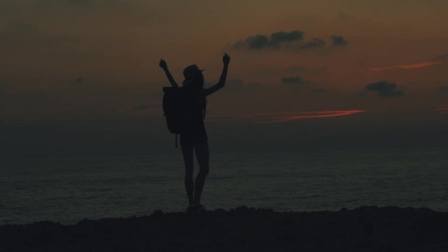 Sexy young traveler woman with a backpack and straw hat on head standing with hands up near the sea on sunset and dawn in Cyprus, turns around and go to the camera , silhouette of a girl, 100 frames