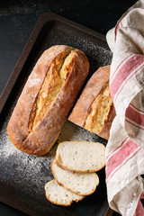 Whole and slice homemade white wheat bread with wheat flour and kitchen towel on old black oven tray as background. Top view