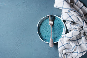 Empty blue ceramic plate with vintage fork and white kitchen towel over blue concrete background. Flat lay with copy space