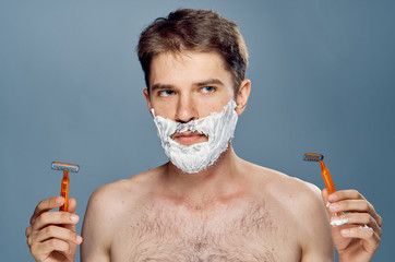 A man with a beard on a gray background holds razors in shaving foam, ortret.emotsii