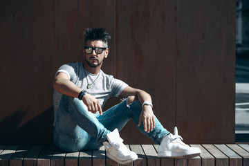 Fototapeta premium Portrait of handsome young man in casual clothing with sunglasses sitting on wood bench, looking away to a side. Color Toned Image. Concept urban man.