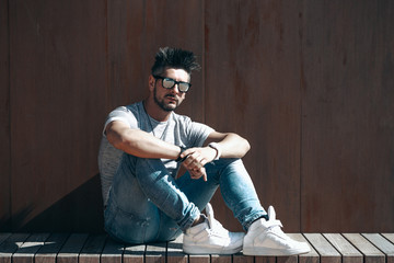 Fototapeta premium Portrait of handsome young man in casual clothing with sunglasses sitting on wood bench, looking away to a side. Color Toned Image. Concept urban man.