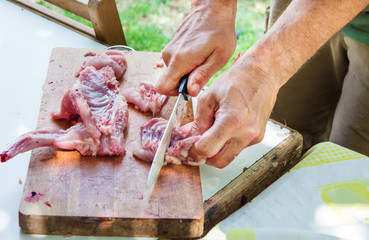 Man's hand holding knife cutting and carving raw rabbit on wood cutting board. Preparing ingredients for paella jambalaya, barbecue. Outdoors, picnic, weekend, nature, summer.