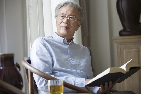 Thoughtful senior man holding book while sitting on chair