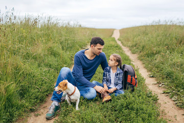 Affectionate daddy sitting near his daughter, having pleasant conversation with her, resting for minute after long walk with their dog. Father, daughter and dog sitting on green path outside