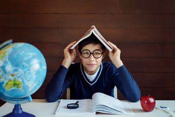 Busy schoolboy, doing his home assigment, keeping book on head, looking puzzled while not knowing...