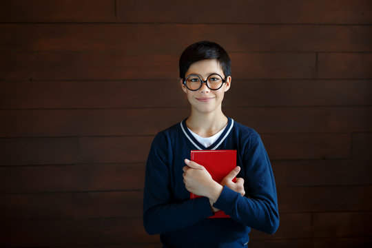 Attractive schoolboy in big glasses and elegant sweater, keeping red book in hands, going to read it. Little mixe race boy wearing spectacles, going to school, posing with red book into camera