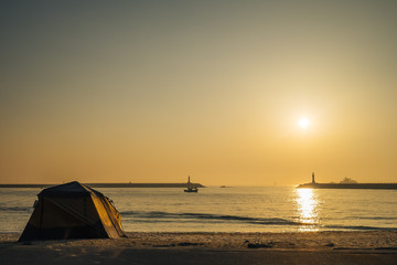 a tent on the beach near by the sea during the sun rise. The sun is shining making the water turn gold. a boat is running out to a deep sea to catch fish. Rock dam protecting the beach and the port. 