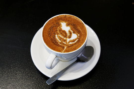 A white cup of cappuccino coffee with a pattern from cinnamon on crema on black table