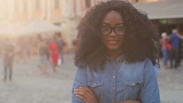 Portrait of beatiful curly hair african woman in denim shirt with crossing hands, smiling on the camera on the old city background.