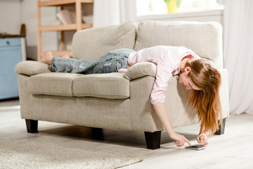 Young girl lying on sofa, typing on smartphone. Female on couch looking on floor, playing with phone.