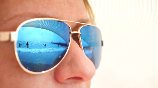 Close-up face of a happy woman in sunglasses. The beach with the resting people is reflected in the glasses. 4k, slow motion
