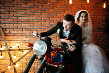 Obraz na płótnie Canvas Amazing wedding couple posing with a huge old motorcycle in a room full of lights.