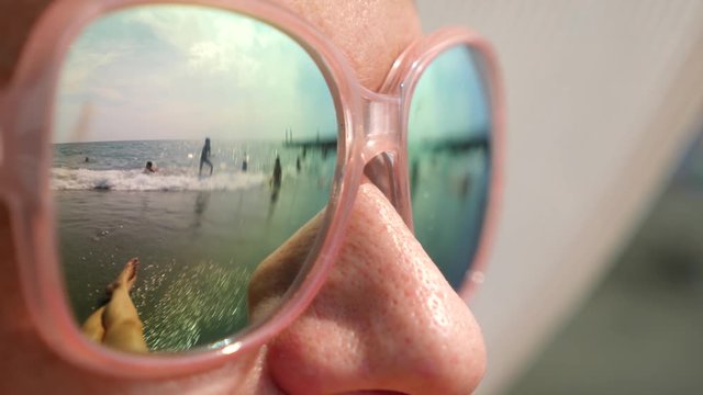 Close-up face of a happy woman in sunglasses. The beach with the resting people is reflected in the glasses. 4k, slow motion