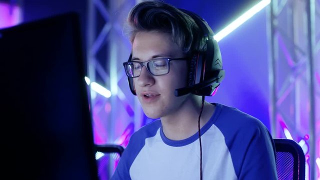 Young Teenage Boy Gamer Plays Video Game on a eSports Tournament/ Internet Cafe. He Wears Glasses and Headphones and Speaks into Microphone. Shot on RED EPIC-W 8K Helium Cinema Camera.