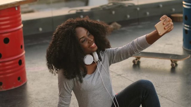 Portrait of young african woman with headphone on the neck using smart phone for making selfie while she sitting on the roof.