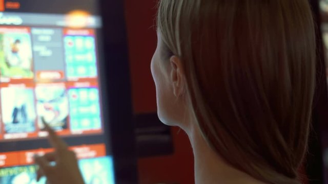 Young pretty woman in red t-shirt choosing movie and buying ticket from vending machine at movie theater