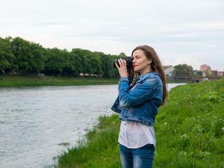 Beautiful girl tourist in a cotton jacket taking photos with a professional camera on the banks of the river in windy weather