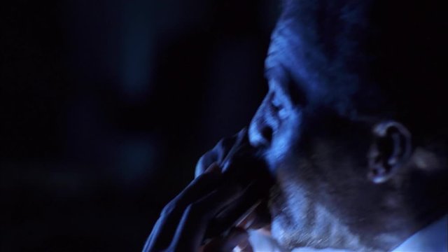 Close-up side shot of blues musician playing harmonica under blue light.
