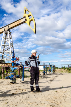 Woman engineer in the oilfield talking on the radio wearing white helmet and work clothes. Industrial site background. Oil and gas concept.
