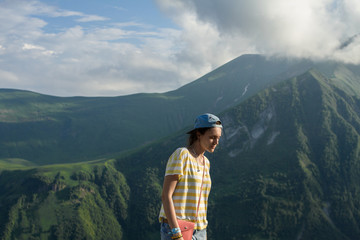 A young girl in a striped yellow T-shirt and a cap in the summer on the fairy-tale mountains and a mysterious misty sky background.