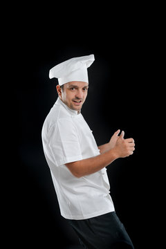 Chef Wearing Uniform With Black Pants And White Hat Isolated On A Black Background Smiling 