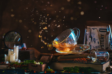 Tea splash in a glass cup on a wooden background with candles, mystery newspaper clips, books,...