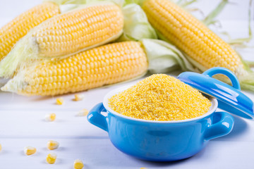 Corn grits polenta in a blue ceramic bowl on white table with ripe raw corn cob and green leaves