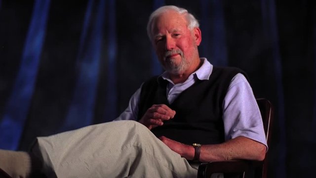 Elderly white man with goatee crosses legs while sitting.
