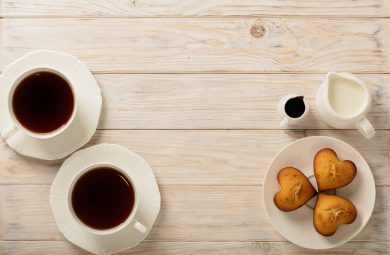 Cups with tea and cupcakes in the shape of a heart on a light wooden background.