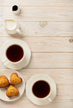 Cups with tea and cupcakes in the shape of a heart on a light wooden background.